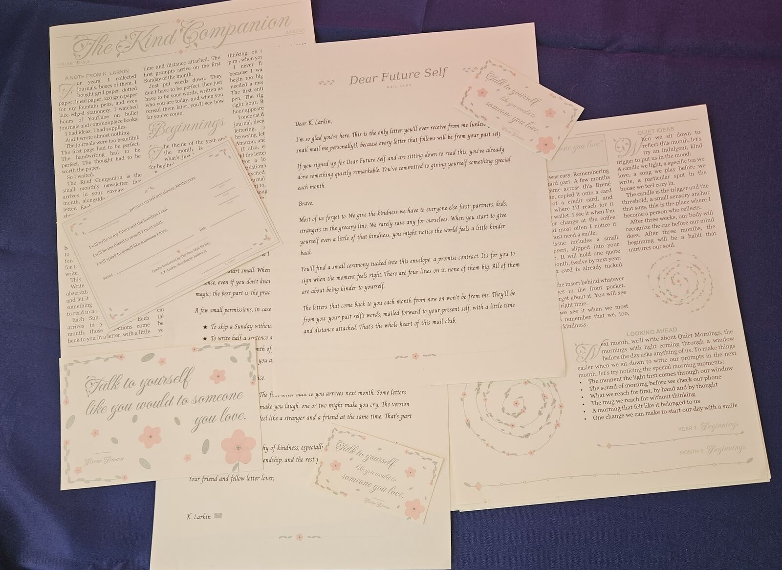 The contents of one monthly Dear Future Self envelope fanned out: the welcome letter, The Kind Companion newsletter, the Promise Contract, the Borrowed Wisdom postcard, and a wallet quote card.