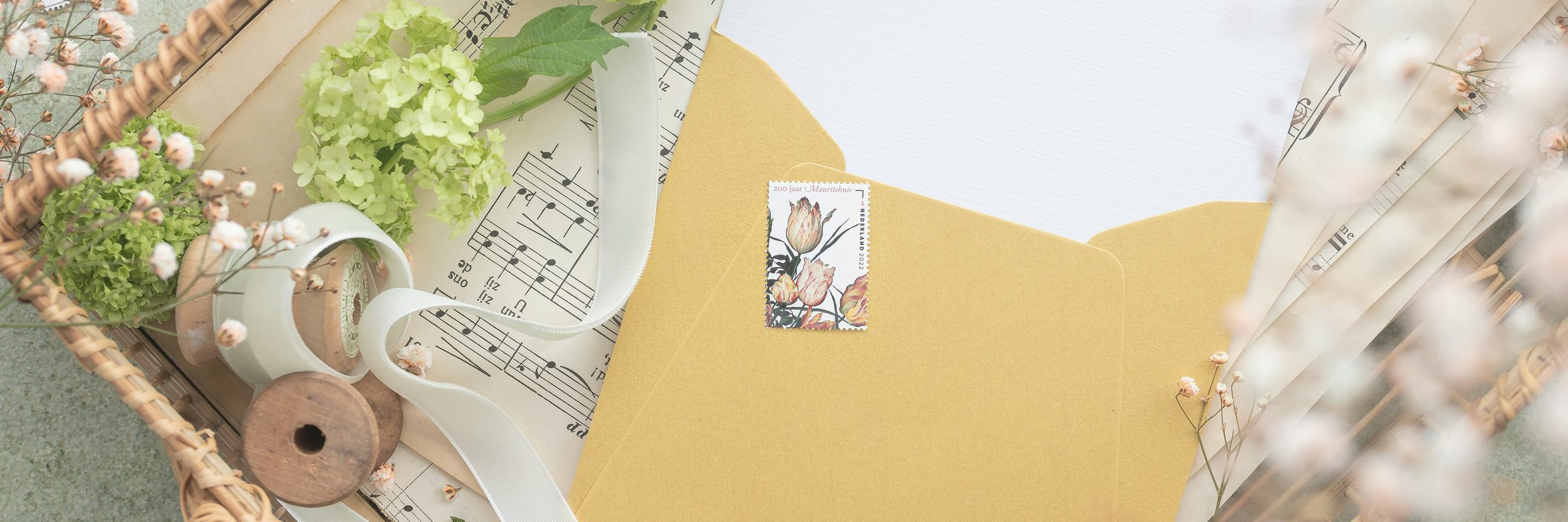 A mustard-yellow envelope resting on sheet music, surrounded by dried flowers, stamps, and ribbon.